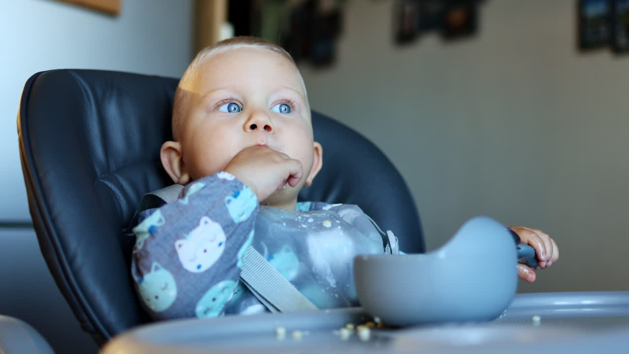 One-year-old boy sits in a high chair and learns to eat by himself. Silicone tableware for children. Boy eats from a plate by himself