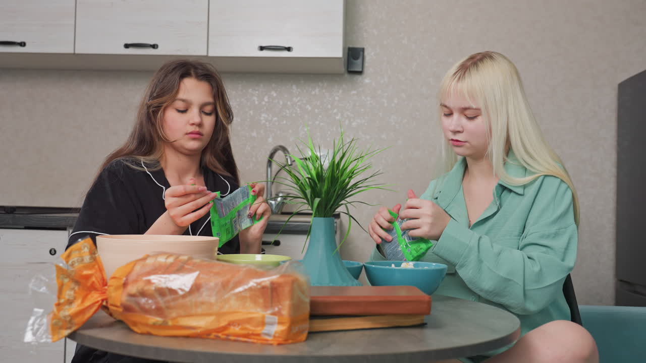 two sisters sitting at round kitchen table with bread loaf on table as one sister pours snacks from green pack into bowl, casual home atmosphere, natural light, relaxed family moment