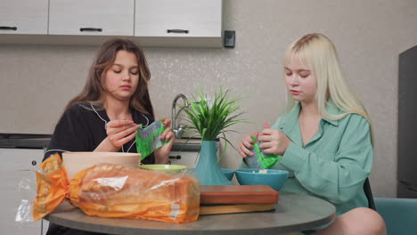 two sisters sitting at round kitchen table with bread loaf on table as one sister pours snacks from green pack into bowl, casual home atmosphere, natural light, relaxed family moment