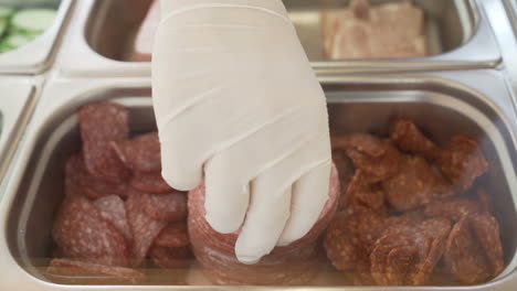 hombre con guantes tomando una rodaja de pepperoni de un recipiente de metal en un bar de sándwiches
