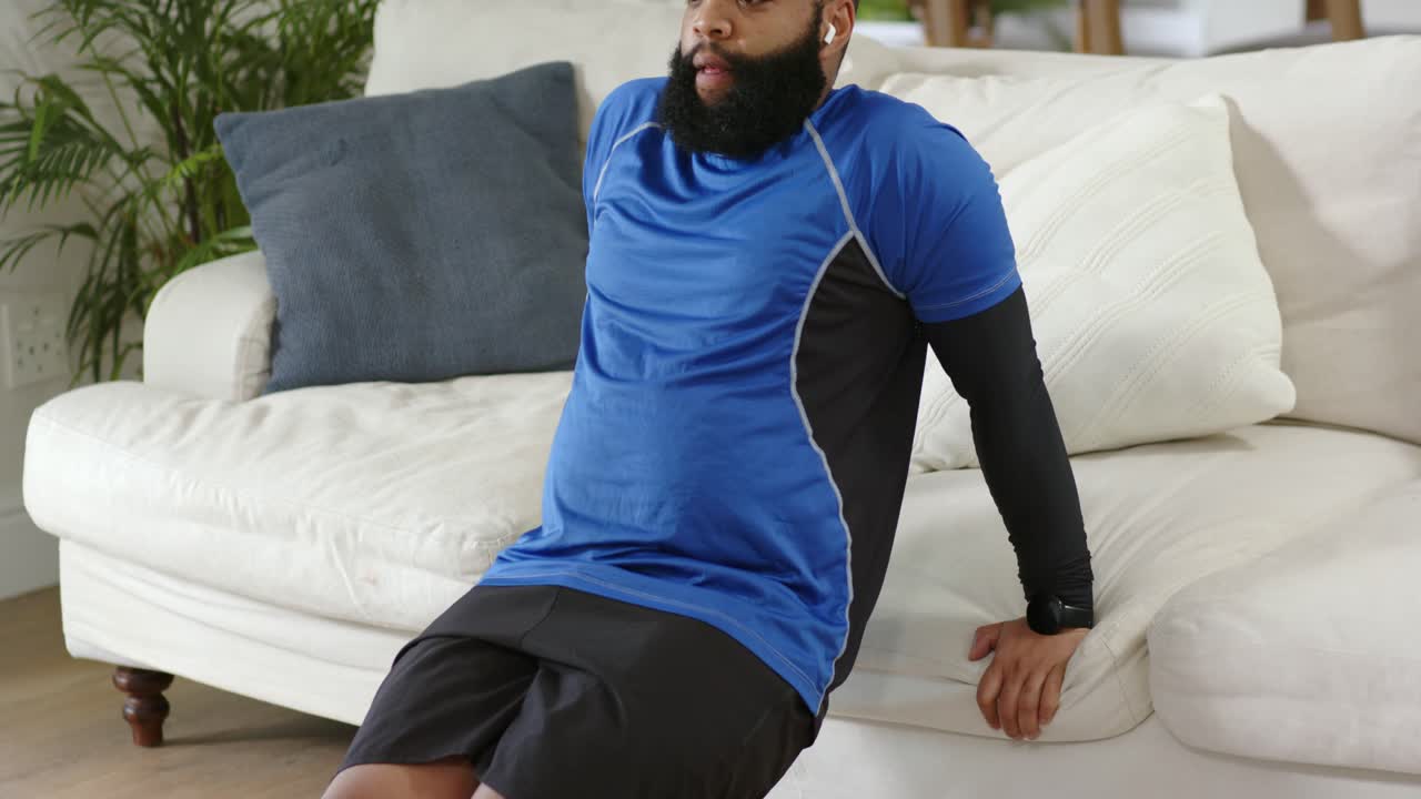 African American man bracing on sofa at home doing dips lowering, rising in shorts for strength