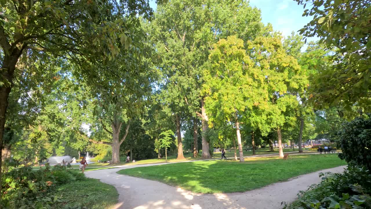 Camera moves through brick gate into green park with trees, winding path, and bright daylight