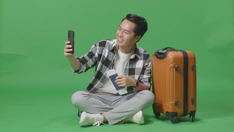 full body of asian male traveler with luggage and passport having video call on smartphone while sitting in the green screen background studio