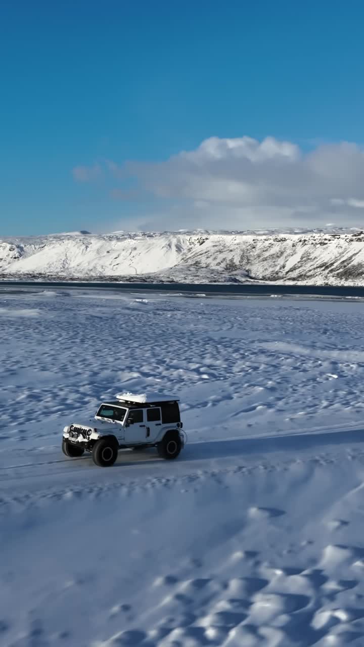 Veduta aerea verticale di un veicolo fuoristrada che attraversa la costa ghiacciata di Kleifarvatn in Islanda, con montagne vulcaniche innevate e un cielo invernale luminoso.