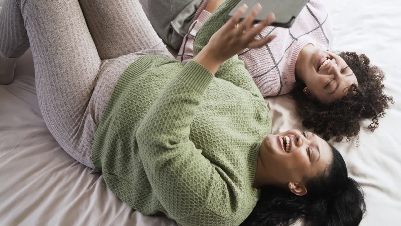 Madre e figlia sdraiate sul letto vedono un tablet alzarsi sopra i loro volti e ridono guardando un video divertente.