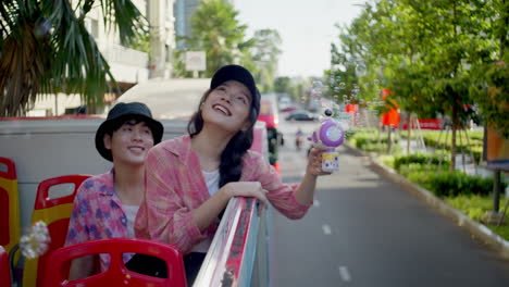 young asiatic couple play with a soap bubble maker toy gun while travelling on open bus in new city during their summer holiday break