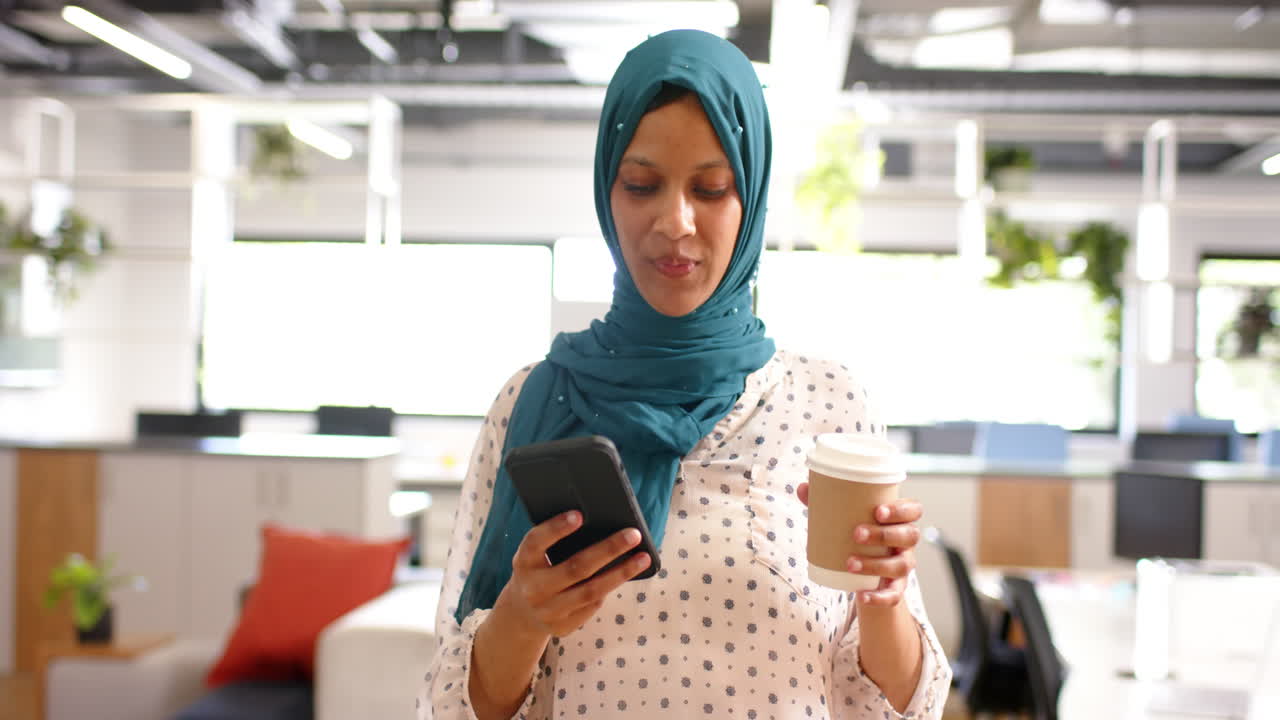 una mujer de negocios de dos razas en hijab bebiendo café y usando un teléfono inteligente en la oficina, en cámara lenta