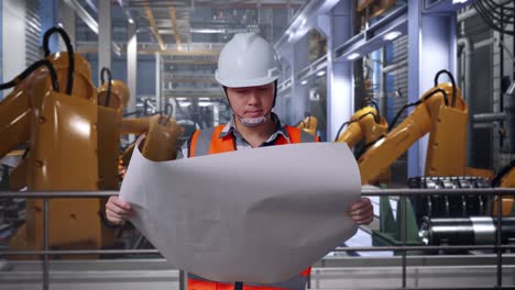 asian male engineer with safety helmet looking at blueprint in his hands and looking around while standing with industrial robotic arms working