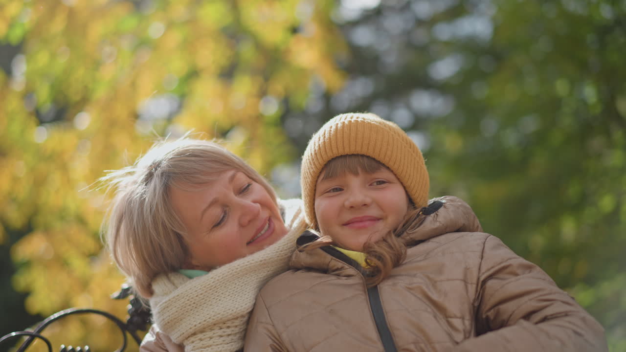 mother holds daughter affectionately as she smiles amid sunlit autumn leaves swaying gently in breeze on park bench, both wearing pastel coats and knit scarf capturing warm family bond