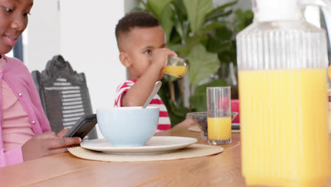 African-american-brother-eating-and-sister-using-smatphone-in-kitchen,-slow-motion