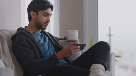 Un-Joven-Feliz-Usando-Un-Teléfono-Inteligente-Navegando-En-Línea-Leyendo-Mensajes-En-Las-Redes-Sociales-Disfrutando-De-Un-Estilo-De-Vida-Cómodo-Bebiendo-Café-Relajándose-En-El-Sofá-De-Casa