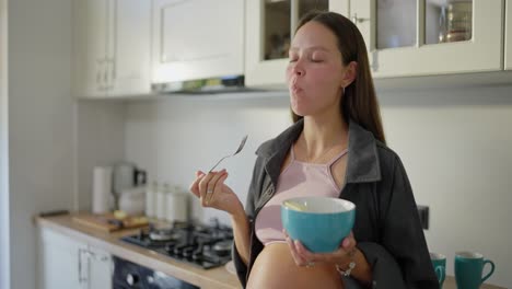 happy pregnant brunette woman with blue eyes eats delicious fruit salad in the kitchen during breakfast in the morning