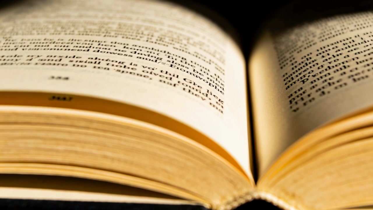 Libro tascabile aperto che si gira su una superficie scura, una leggera brezza che solleva una pagina, luce calda che rivela il testo.