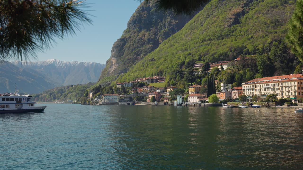 Una vista panoramica sul Lago di Como rivela un incantevole villaggio italiano con ville colorate che scendono a cascata lungo rigogliose colline verdi fino al lungolago con lontane cime alpine innevate nella luce primaverile