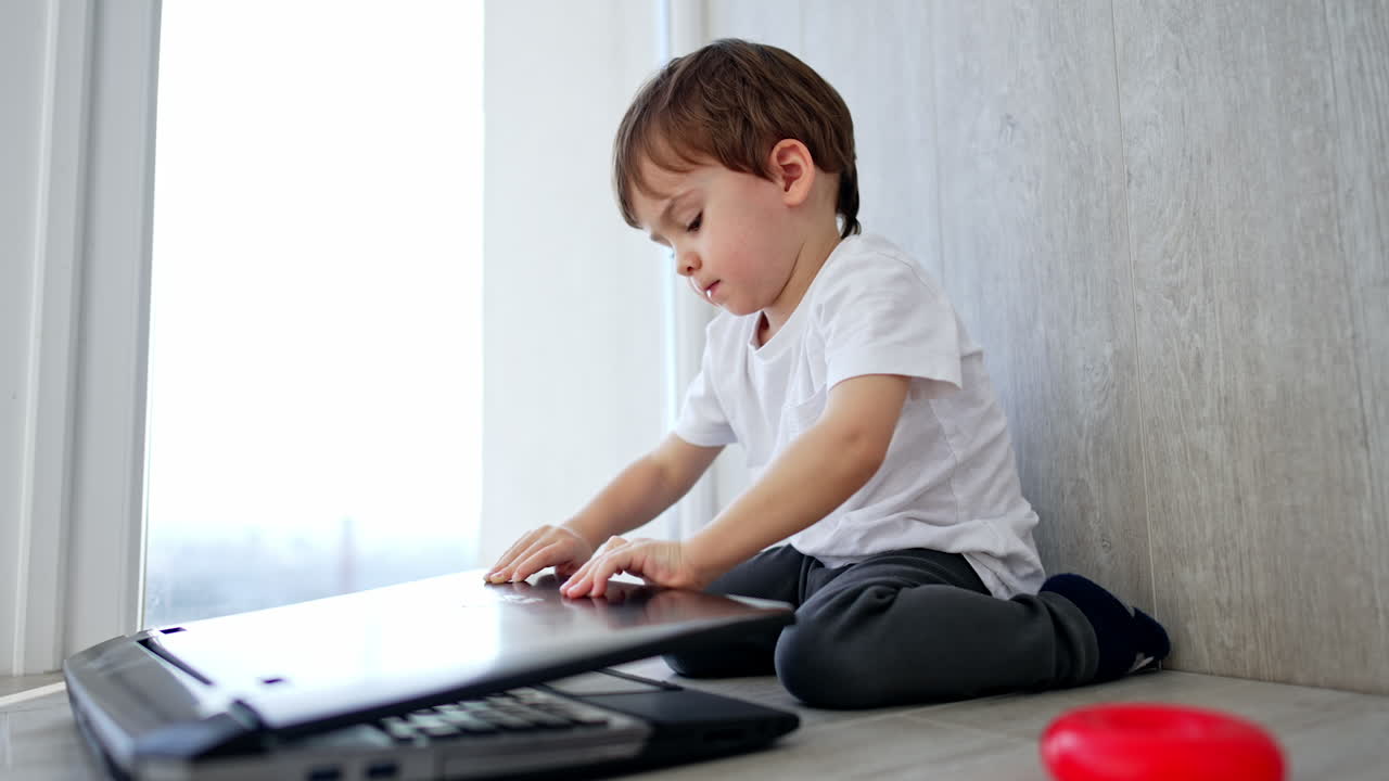 Lovely kid opens a laptop on the floor in front of him. Toddler looks at gadget smiling. Low angle view.