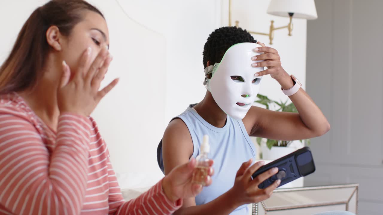 Diverse Freundinnen auf dem Bett, links Serum auftragend, rechts LED-Maske anlegend, tippen auf Fernbedienung für die Hautpflege.