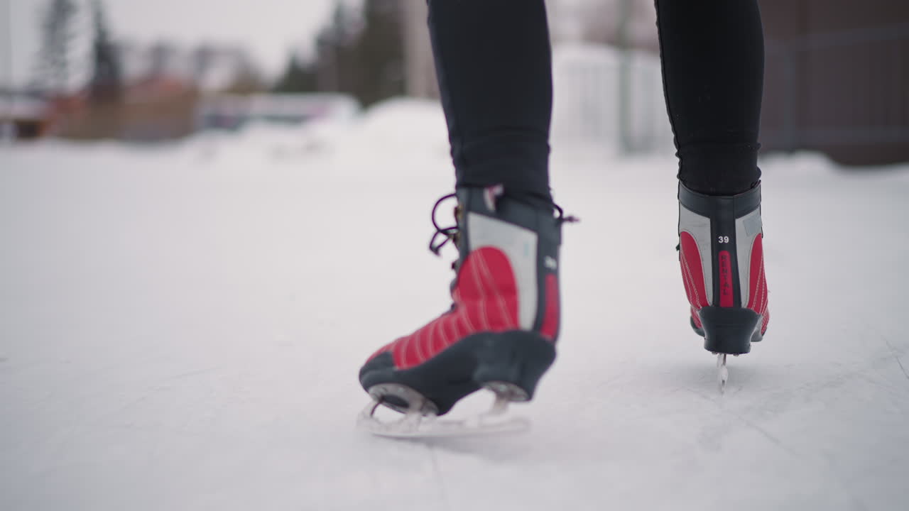Nahaufnahme einer Person mit roten Schlittschuhen, die im Winter auf einer Eisbahn im Freien gleitet, Beine in schwarzen Hosen, die sich mit Gleichgewicht und Konzentration bewegen, Schnee und verschwommene Gebäude im Hintergrund, kalte saisonale Atmosphäre