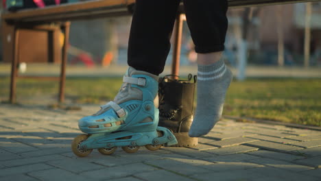 A person sitting in a park, with one leg in a rollerblade, pulling off a black boot from the other leg and preparing to wear the second rollerblade