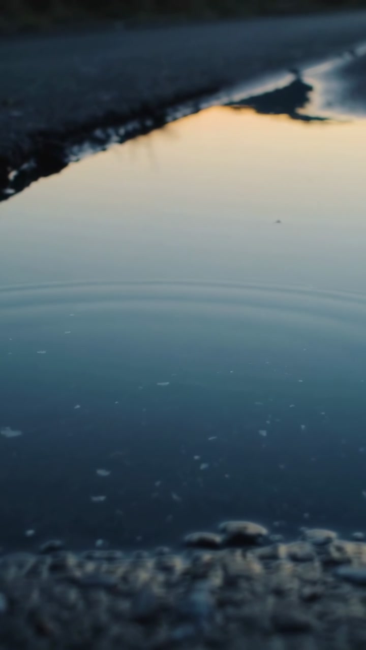 Vertical video: Rippling road puddle showing ripples spreading and calming after droplet at sunset