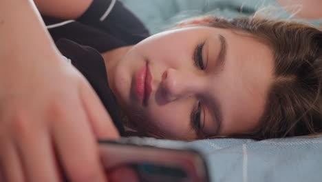 Young girl lying sideways on bed holding smartphone close to face while staring at screen with calm focused expression, cozy setting suggests relaxed moment during leisure