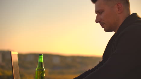 Young-male-freelancer-sits-on-the-roof-with-a-laptop-and-beer-and-types-on-the-keyboard.