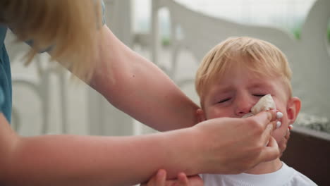 a young boy struggles as his mother gently wipes his mouth clean with a tissue