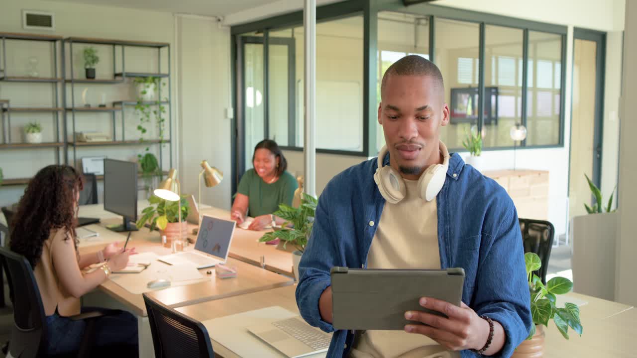 African man sitting on desk holding tablet, tapping and scrolling, reacting to colleague laugh