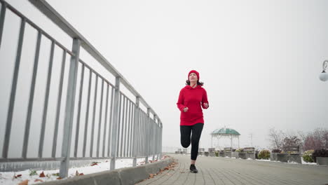 woman jogging in red beanie and jacket along snowy pathway near iron railing in foggy atmosphere, decorative structure, light pole, and benches in background, showcasing determination