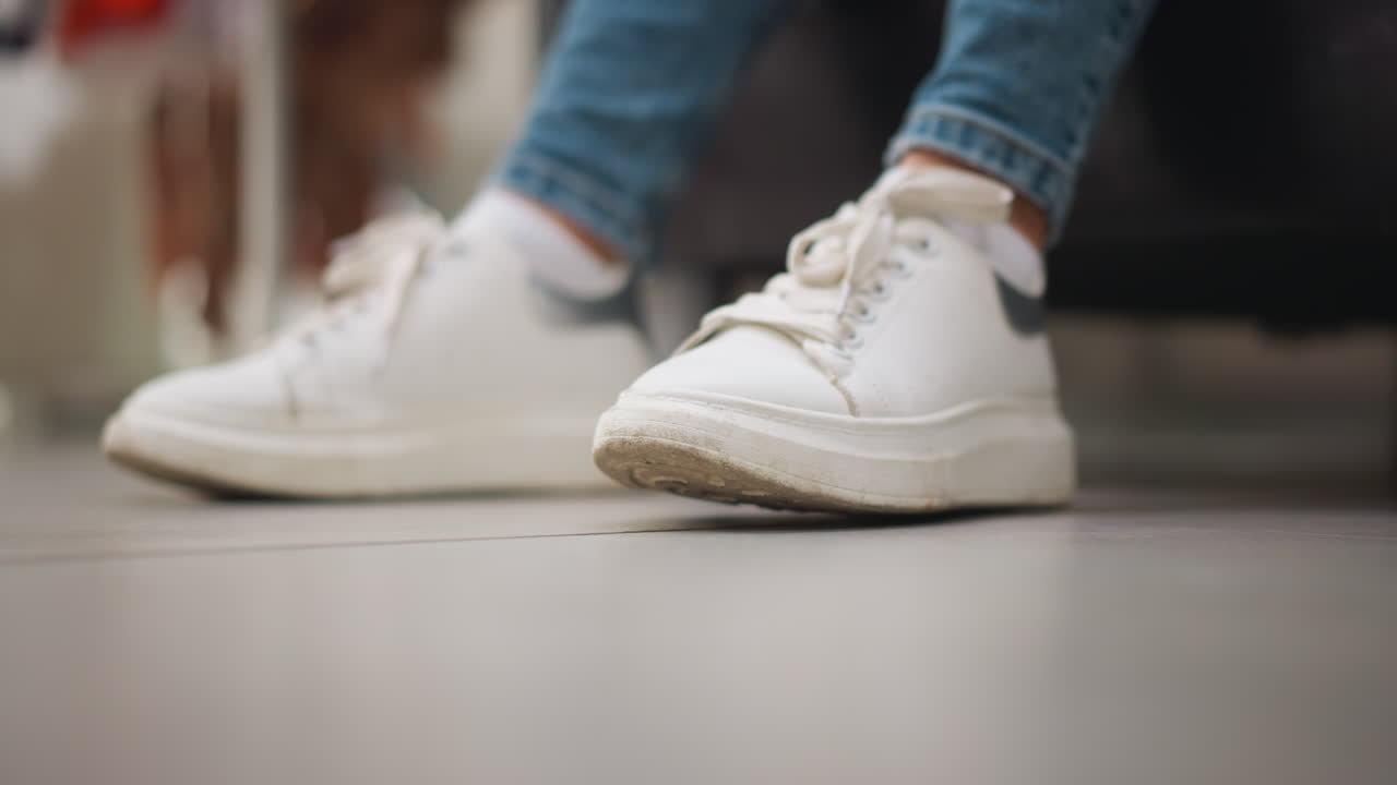 여성의 색 캔버스 스니커의 클로즈업, 부드러운 조명과 함께 쇼핑몰에서 편안한 캐주얼 분위기, 신발에 초점을 맞춘 청바지 바지와 함께 부드럽게 바닥을 두드립니다.