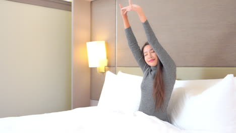 pretty asian woman with long hair wearing grey top stretches arms over head with eyes closed sitting in bed with comfortable puffy white linen and pillows