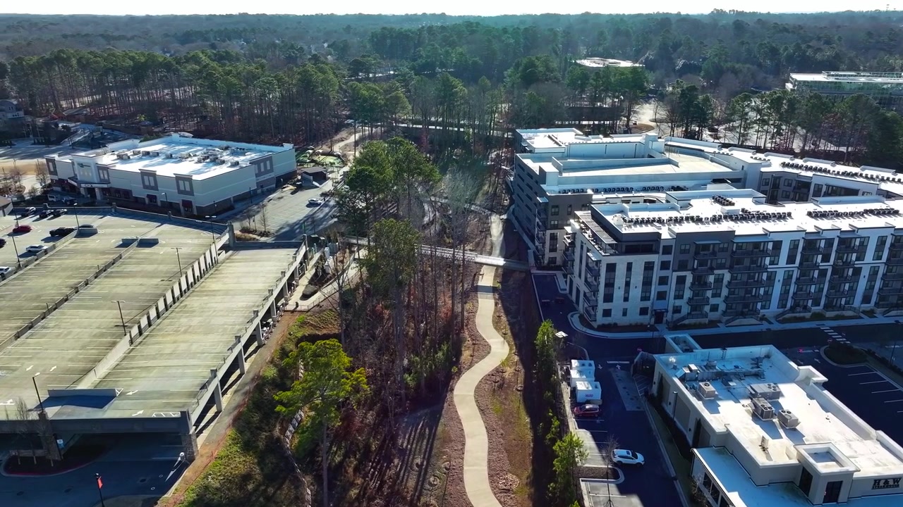 Các khu chung cư hiện đại và tòa nhà bán lẻ ở Atlanta, Georgia với bãi đỗ xe nhiều tầng và lối đi bộ nép mình giữa những cây thông xanh dưới bầu trời nắng chói chang vào ban ngày. Cảnh quay quỹ đạo từ trên không