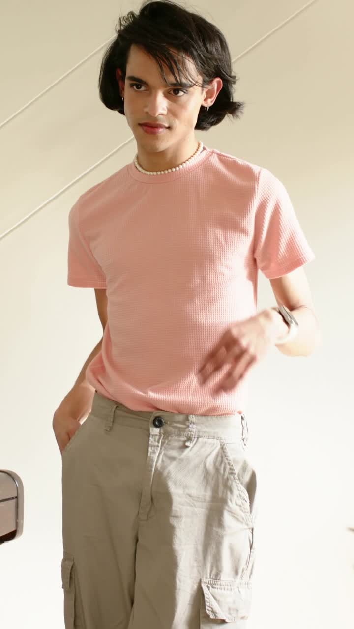 Video verticale: Uomo in maglietta rosa in un loft che registra, alza il mento, sistema i capelli e posa con un orologio bianco.