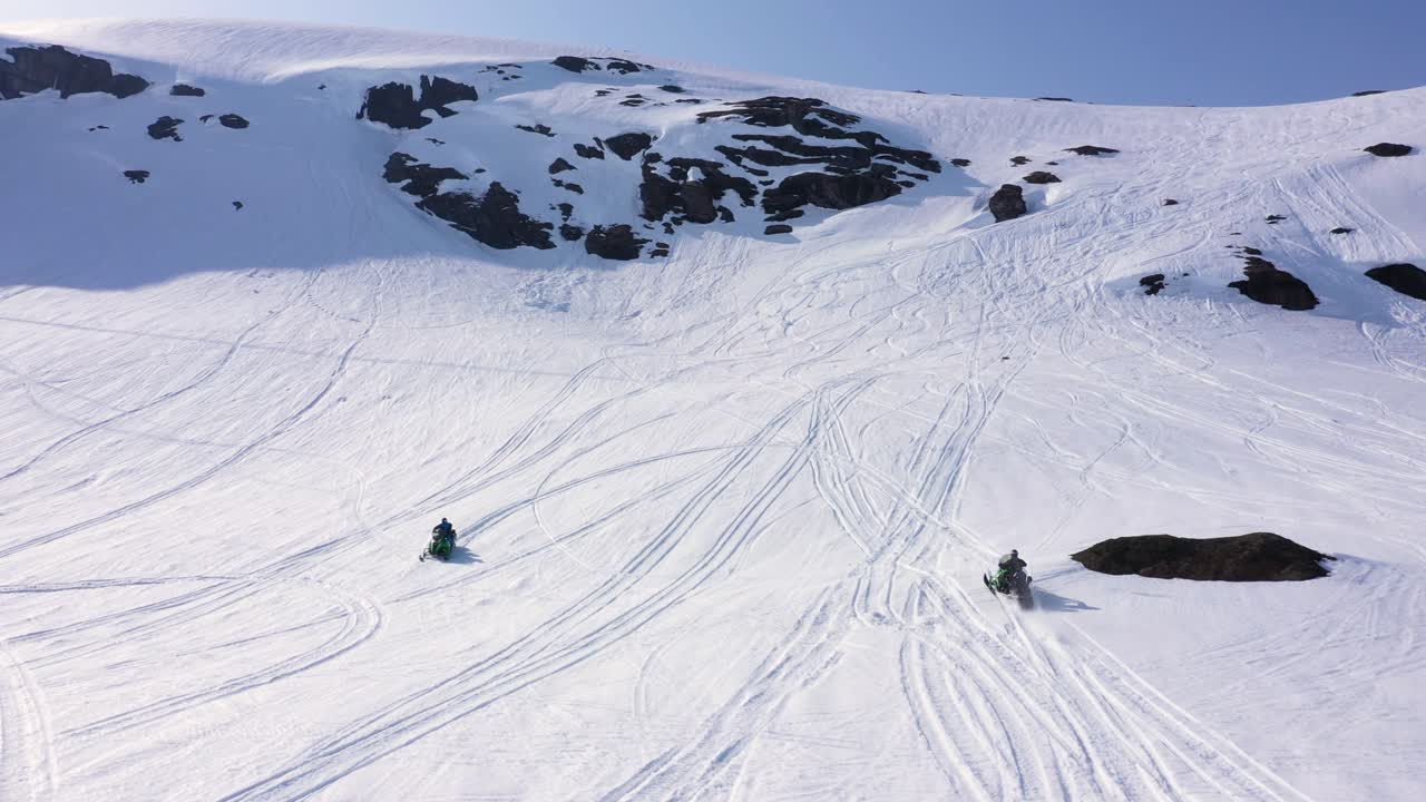 Aerial view over snowy mountain landscape. Snowmobiles driving up mountain side. Sunny weather. MOVING FORWARD, PAN UP.