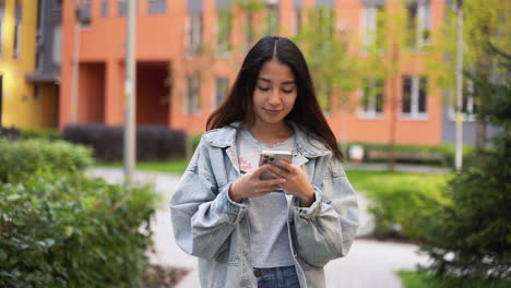 Hermosa-Joven-Japonesa-Enviando-Mensajes-De-Texto-En-El-Teléfono-Móvil-Mientras-Camina-Al-Aire-Libre