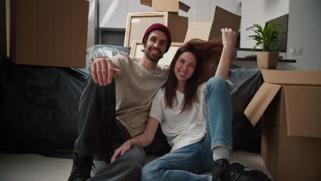 Portrait-of-a-happy-brunette-man-with-stubble-in-a-red-hat-and-a-beige-T-shirt-who-sits-on-the-floor-with-his-brunette-girlfriend-in-a-white-T-shirt-near-the-sofa-in-a-black-cellophane-case-among-a-pile-of-boxes-in-a-new-house-after-a-housewarming-party