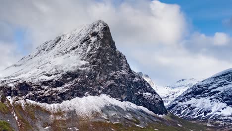 a solitary mount hereggja towering above lush herdalen valley. the huge dark rock sprinkled with fresh snow. thin white clouds cover the bright blue sky, whirling around the mountain top.