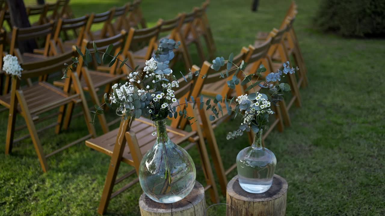 Glass vases with delicate flowers line wooden stumps beside folding chairs for a wedding