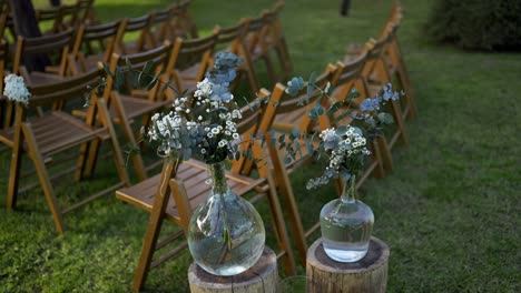 glass vases with delicate flowers line wooden stumps beside folding chairs for a wedding