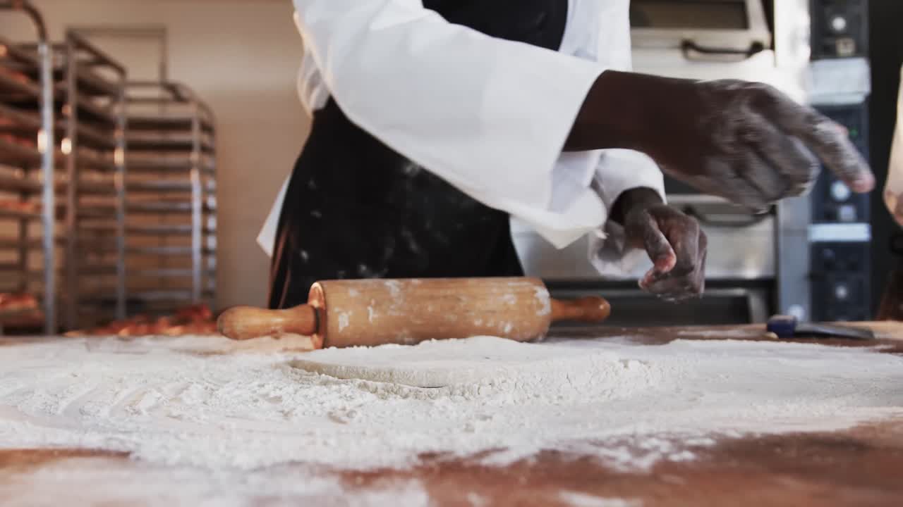 African american male baker working in bakery kitchen, rolling dough on counter in slow motion