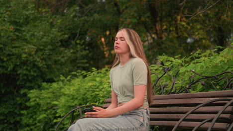 young woman outdoors, holding phone in her right hand and glancing away thoughtfully and sits on the wooden bench, wearing casual outfit with crop top and jeans, surrounded by lush greenery