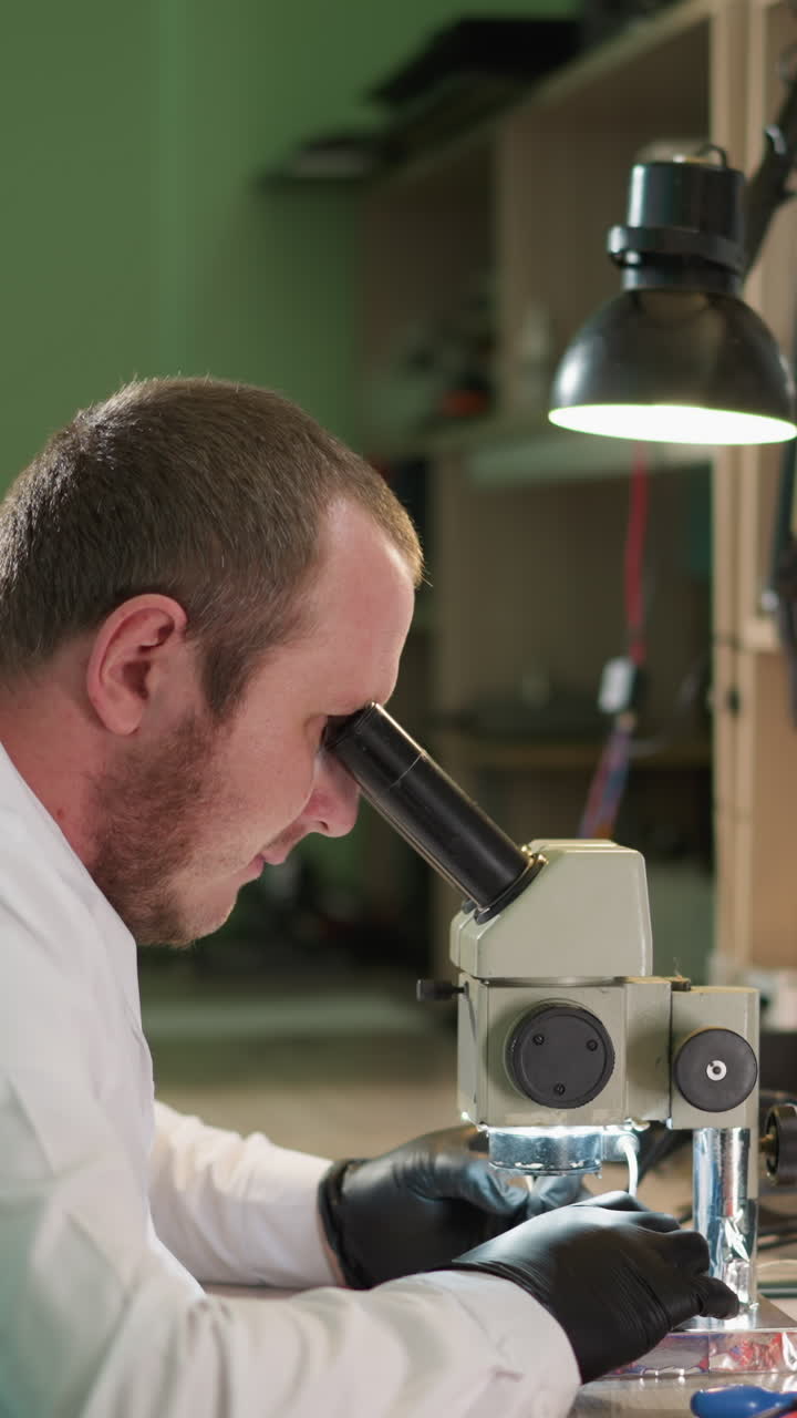 A close-up view of a technician meticulously working on a circuit under a microscope in a laboratory, then relaxing back, the workspace is equipped with a desk lamp and various tools