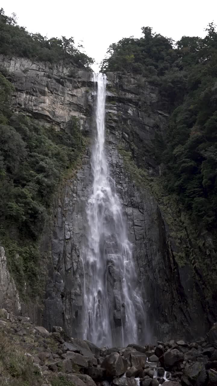 Breathtaking view of Nachi Falls flowing over rugged cliffs surrounded by greenery. Vertical Video, Slow Motion