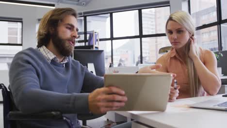 Disabled-caucasian-businessman-sitting-on-wheelchair-using-digital-tablet-talking-to-coworker-in-off