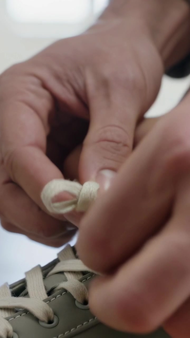 Video verticale: Mani adulte che allacciano una sneaker color oliva con lacci color crema, a casa, con un polsino grigio in vista, in preparazione.