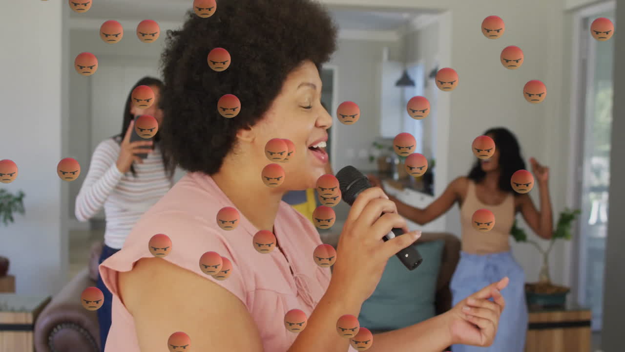 Mid-adult woman singing into microphone at home, friends dancing with angry emojis for social media