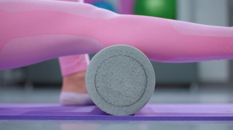 Close-up of legs using a foam roller during a fitness session on yoga mats in a well-lit gym, emphasizing stretching and muscle recovery techniques