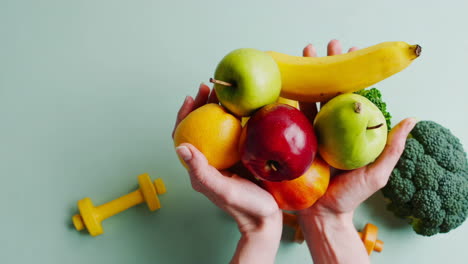 hands holding a variety of fresh fruits and vegetables alongside fitness equipment