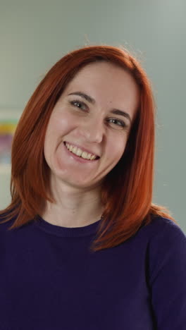 young woman with ginger hair looks in camera tilting head to side. portrait of positive female with smile on face standing against lamp in room closeup