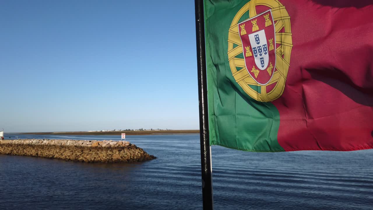 Bandeira portuguesa tremula em primeiro plano, marina de Olhão ao fundo, Portugal, close-up extremo