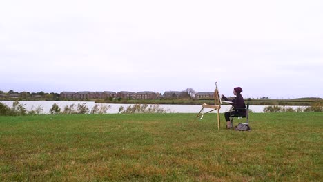 Young-Woman-Pints-Painting-Next-To-Park-Lake-on-Cloudy-Day-Wide-Shot